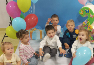 Dzieci siedzą na dywanie i pozują do urodzinowego zdjęcia za nimi jest urodzinowe tło i kolorowe balony.