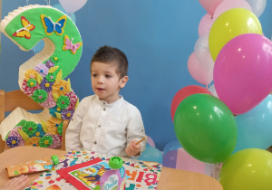 Lavrentii siedzi przy stole pozuje do urodzinowego zdjęcia z tyłu jest tło z balonami i cyfra trzy oraz stojak z kolorowymi balonami.