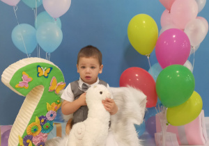 Tadej pozuje do urodzinowego zdjęcia z cyfrą dwa oraz z kolorowymi balonami, w ręku trzyma pluszową maskotkę.