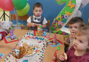 Tadej, Szymon i Aurelia siedzą przy stole, jedzą urodzinowy poczęstunek, za nimi jest urodzinowe tło, kolorowe balony i cyfra dwa.
